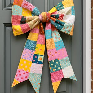 May include: A large, colorful fabric bow made of patchwork squares, displayed on a gray door. The bow features a variety of patterns and colors, including pink, yellow, orange, and teal. The bow is a decorative item.