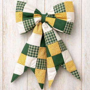 May include: A large, decorative bow made from quilted fabric. The bow has a patchwork design with squares of green plaid, yellow floral, and off-white fabric. The bow is tied in the centre and has long, pointed tails.