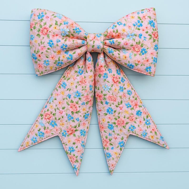 May include: A large, decorative bow with a floral pattern. The bow features a light pink background with a repeating pattern of pink, blue, and yellow flowers. The bow is made of fabric and has a stitched edge.