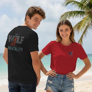 Peut inclure: Deux personnes portent des t-shirts avec des motifs graphiques. L'une porte un t-shirt noir avec "THE WOLF MENTALITY" dans une police stylisée. L'autre porte un t-shirt rouge avec un motif plus petit. Les deux chemises sont à manches courtes.