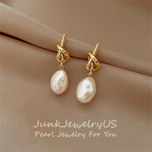 Puede incluir: Un par de pendientes dorados con diseño de nudo y perlas blancas en forma de lágrima. Los pendientes cuelgan de una tela color beige. El texto "JunkJewelryUS Pearl Jewelry For You" está en la parte inferior.
