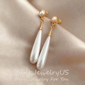 Könnte beinhalten: Ein Paar goldfarbene Ohrringe mit weißen, tropfenförmigen Perlen. Die Ohrringe haben eine kleine Perle oben und einen langen, glatten Perlenanhänger. Der Hintergrund ist ein weicher, neutraler Stoff. Der Text "Junk JewelryUS Pearl Jewelry For You" befindet sich unten.