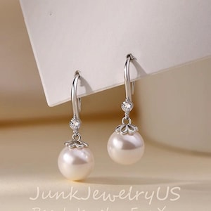 Puede incluir: Elegantes pendientes colgantes plateados. Cada pendiente presenta una gran perla blanca redonda suspendida de un delicado gancho. Una pequeña piedra transparente y brillante adorna el gancho y el engaste floral de la perla. Fondo neutro.