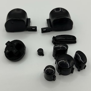 Könnte beinhalten: Eine Sammlung schwarzer Ersatzknöpfe und -komponenten für einen Gamecontroller. Das Bild zeigt verschiedene Formen, darunter D-Pads, Thumbsticks und Schultertasten, alle in glänzend schwarzer Ausführung. Die Teile sind auf einer weißen Oberfläche angeordnet.