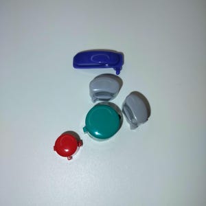 Könnte beinhalten: Eine Auswahl an farbenfrohen Kunststoff-Spielkonsolenknöpfen und -komponenten. Die Sammlung umfasst einen dunkelblauen Knopf, zwei graue Knöpfe, einen teal-farbenen Knopf und einen roten Knopf. Die Artikel sind auf einer weißen Oberfläche angeordnet.