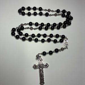 May include: A black rosary with round beads and a silver crucifix. The rosary features a detailed silver cross with a figure of Jesus and a small medal. The beads are strung on a silver chain.