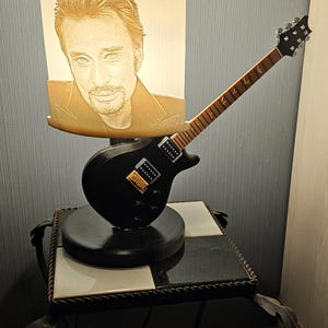 Peut inclure: Lampe en forme de guitare électrique noire avec un portrait d'homme sur l'abat-jour. La guitare a un micro doré et un manche marron. La lampe est posée sur une petite table à plateau noir et blanc.