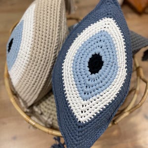 May include: Two crochet eye-shaped pillows in a wicker basket. One pillow is navy blue with a light blue and white eye design, and a tassel. The other pillow is beige with a light blue and white eye design.
