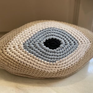 May include: Crocheted eye-shaped pillow in beige, white, light blue, and black. The pillow features concentric circles of color, resembling an eye. The handmade pillow is a decorative accent for a bed or sofa.
