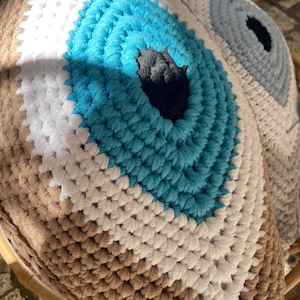 Op de afbeelding: Gehaakte kussens in de vorm van een oog, in bruin-, wit- en turkoois tinten. De kussens hebben een centraal oogontwerp met een zwarte pupil. Het getextureerde haken creëert een unieke, handgemaakte esthetiek, ideaal voor woondecoratie.