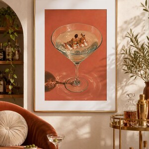 May include: A framed wall art print featuring a large martini glass filled with clear liquid against a solid terracotta background. Inside the glass, two small figures in swimwear lounge on a striped towel floating on the surface of the liquid.
