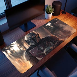 Könnte beinhalten: Ein rechteckiges Mauspad mit einer detaillierten Illustration von The Mandalorian und Grogu aus der Star Wars-Serie. Das Design zeigt die Charaktere in einer Waldkulisse mit warmem, goldenem Licht.