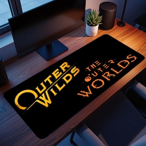 May include: A black desk mat with the words "Outer Wilds" and "The Outer Worlds" in gold. The mat is rectangular and placed on a wooden desk. A computer monitor, speakers, and a small potted plant are visible in the background.