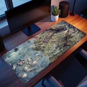 May include: A rectangular desk mat featuring a detailed map design with shades of blue, green, and brown. The map depicts a landscape with mountains, water, and various settlements. The mat is placed on a wooden desk, with a computer monitor and a small potted plant visible.