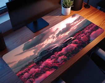 May include: A rectangular mousepad featuring a black car driving through a field of pink flowers under a cloudy sunset sky. The design is printed on a long, rectangular surface, suitable for a desk.