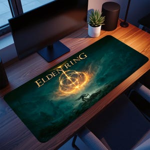 Könnte beinhalten: Rechteckiges Gaming-Mauspad mit dem goldenen Schriftzug "ELDEN RING". Das Design zeigt ein feuriges Emblem und eine Figur in einer dunklen Landschaft. Das Mauspad liegt auf einem Holzschreibtisch mit einem Computermonitor und einer Topfpflanze.