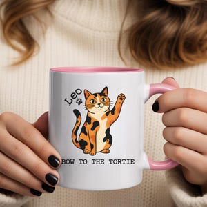 Peut inclure: Mug en céramique blanche avec intérieur et anse roses. La tasse présente une illustration de chat de dessin animé en orange, noir et blanc, avec le texte "Leo" et "BOW TO THE TORTIE".