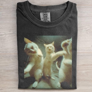 Puede incluir: Camiseta gris oscuro Comfort Colors con una imagen humorística de tres gatos blancos en pose de baile. Los gatos están sobre un fondo apagado. La camiseta tiene un cuello redondo clásico y una etiqueta en la parte superior central.