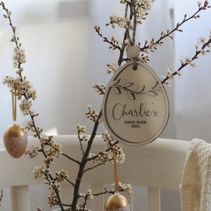 Op de afbeelding: Een Paasdecoratie met bloeiende takken in een witte kruik, versierd met hangende eieren en een houten ei-vormig ornament. Het ornament leest "Charlie's Eerste Pasen 2026". De eieren hebben gouden en gespikkelde roze afwerkingen.