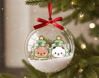 May include: Clear glass Christmas ornament with a red ribbon bow. Inside, two cartoon characters are dressed as Christmas trees, set against a snowy backdrop. The ornament hangs from a green Christmas tree.