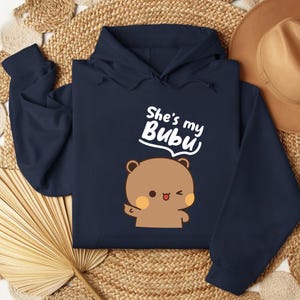 May include: Navy blue hoodie with a cartoon bear illustration and the text "She's my Bubu" in white. The bear has brown fur, orange cheeks, and a winking expression. The hoodie has a drawstring hood and long sleeves.