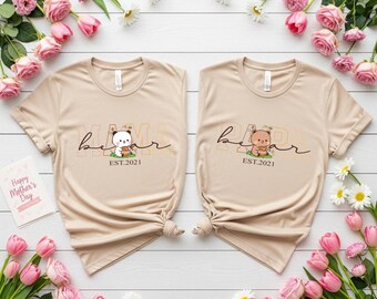 May include: Two beige t-shirts with the word "Mama Bear" in a script font, each featuring a bear graphic and "EST.2021". The shirts are surrounded by pink roses, tulips, and daisies. A "Happy Mother's Day" card is also present.