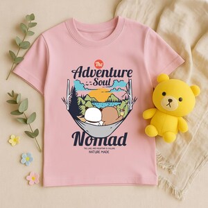 Peut inclure: T-shirt rose clair avec un motif de deux ours dans un hamac, des montagnes et le texte "The Adventure Soul Nomad". Un ours en peluche jaune est &agrave; c&ocirc;t&eacute; du t-shirt. Le texte "THE LAKE AND MOUNTAIN IS CALLING NATURE MADE" est &eacute;galement pr&eacute;sent.