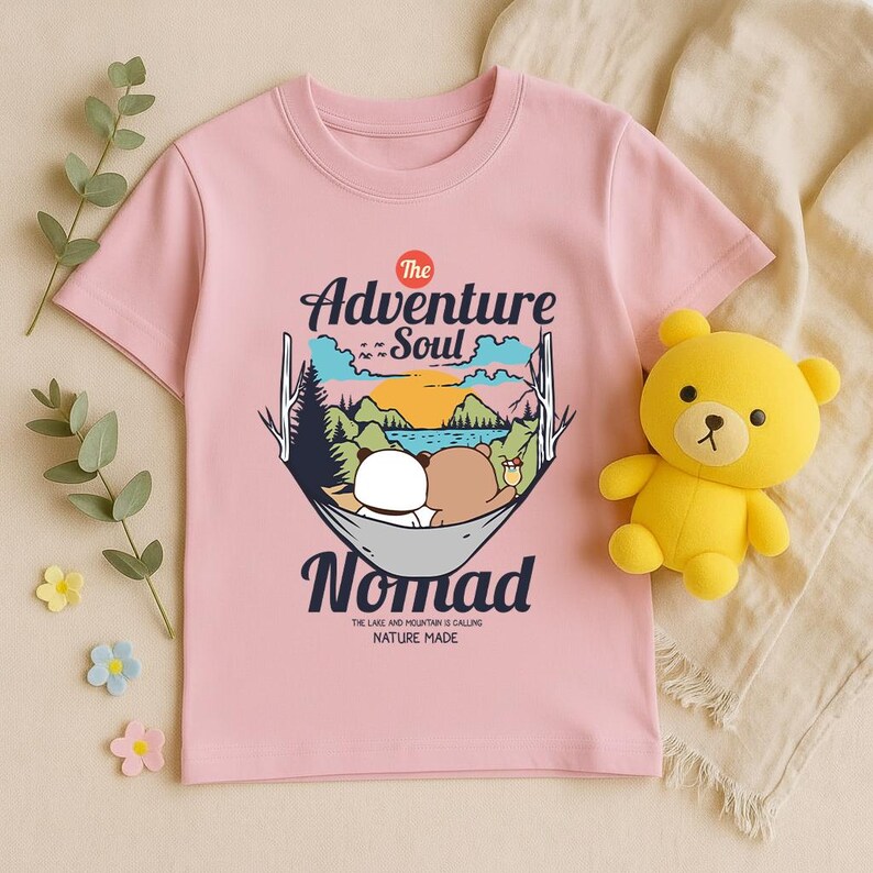 Peut inclure: T-shirt rose clair avec un motif de deux ours dans un hamac, des montagnes et le texte "The Adventure Soul Nomad". Un ours en peluche jaune est &agrave; c&ocirc;t&eacute; du t-shirt. Le texte "THE LAKE AND MOUNTAIN IS CALLING NATURE MADE" est &eacute;galement pr&eacute;sent.