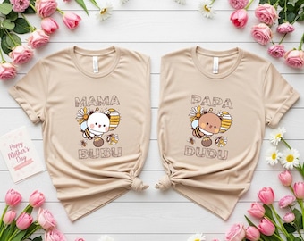May include: Two beige t-shirts with matching designs. One says "Mama Bubu" and the other says "Papa Dudu". Each shirt features a cartoon bee with a flower and honeycomb design. Pink tulips and daisies surround the shirts.