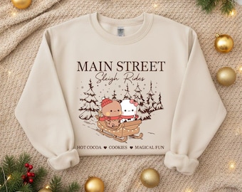 May include: Beige sweatshirt with a winter scene graphic. The graphic features two cartoon characters in a sleigh with the text "MAIN STREET Sleigh Rides". Additional text includes "HOT COCOA COOKIES MAGICAL FUN". Christmas-themed apparel.