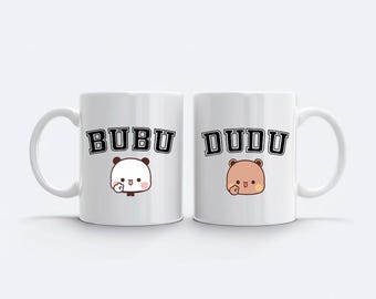 May include: Two white ceramic mugs with the names "BUBU" and "DUDU" printed in black block letters. Each mug features a cartoon animal character. The BUBU mug has a panda, and the DUDU mug has a brown bear.