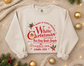 May include: A cream-colored sweatshirt with the text "White Christmas" in red, featuring "The Cozy Bear Couple" and "The Cuddle Inn, Snowville, VT, Winter 1954." The design includes holly and bear illustrations, perfect for the holiday season.