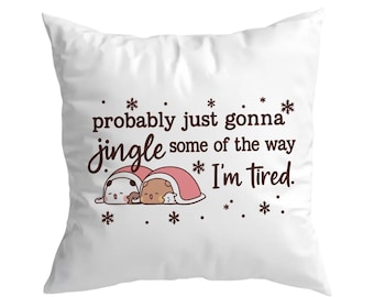 May include: White square pillow with the text "probably just gonna jingle some of the way I'm tired." in brown script. Two cartoon characters wearing Santa hats are under a red blanket. Snowflakes are scattered around the text and characters.