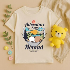 Peut inclure: T-shirt cr&egrave;me avec un motif de deux ours en dessin anim&eacute; dans un hamac et le texte "The Adventure Soul Nomad". Le t-shirt porte &eacute;galement l'inscription "The Lake and Mountain is Calling Nature Made". Un ours en peluche jaune est &agrave; droite.