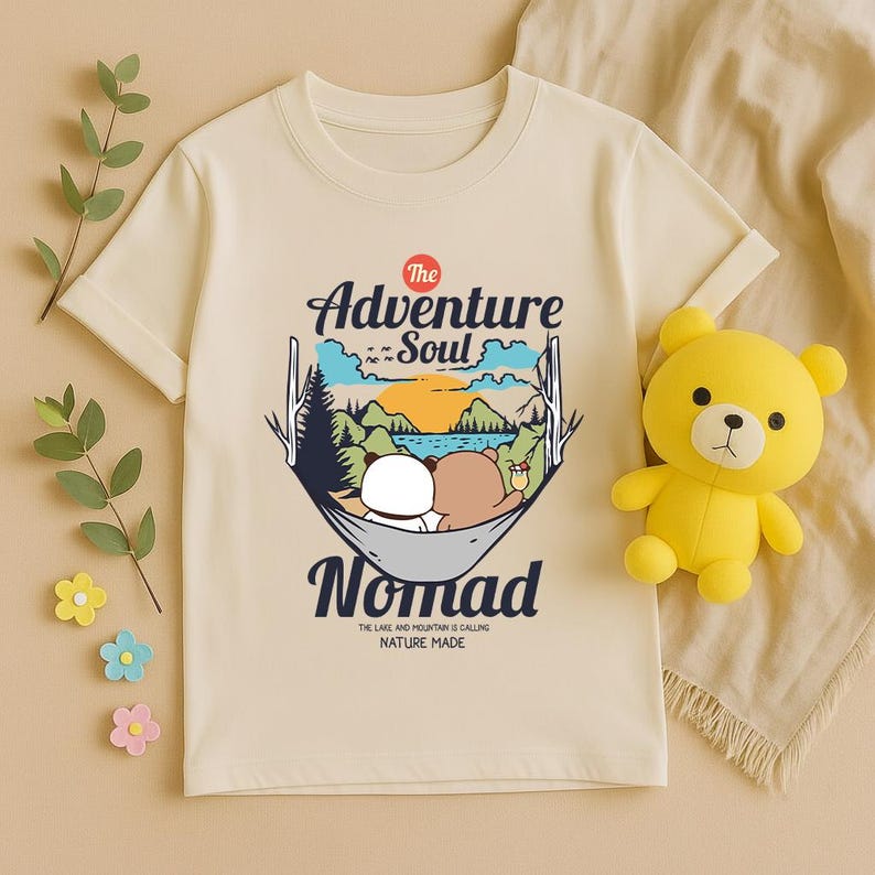 Peut inclure: T-shirt cr&egrave;me avec un motif de deux ours en dessin anim&eacute; dans un hamac et le texte "The Adventure Soul Nomad". Le t-shirt porte &eacute;galement l'inscription "The Lake and Mountain is Calling Nature Made". Un ours en peluche jaune est &agrave; droite.