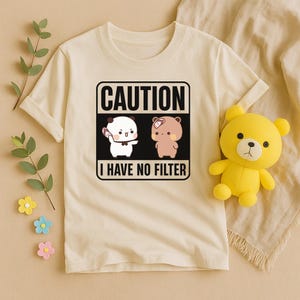 May include: Cream-colored t-shirt with a graphic that reads "CAUTION I HAVE NO FILTER." The graphic features two cartoon characters. A yellow teddy bear toy is next to the shirt. The shirt has short sleeves and a crew neck.