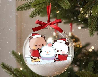 May include: Clear Christmas ornament with a red ribbon, featuring a cartoon bear and panda in Santa hats, building a snowman. The ornament is filled with faux snow and is hanging on a Christmas tree.