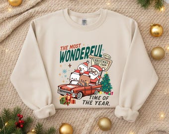 May include: Beige sweatshirt with a Christmas-themed graphic. The graphic features a red car carrying Santa Claus, a snowman, and a bear, with the text "THE MOST WONDERFUL TIME OF THE YEAR."