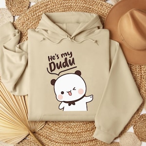 May include: A beige hoodie with the text "He's my Dudu" above a cartoon panda bear. The panda has black ears, a black bow tie, and is winking. The hoodie has a drawstring hood.