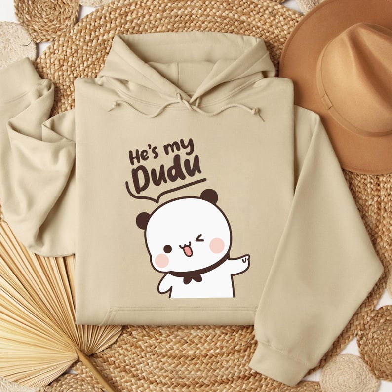 May include: A beige hoodie with the text "He's my Dudu" above a cartoon panda bear. The panda has black ears, a black bow tie, and is winking. The hoodie has a drawstring hood.