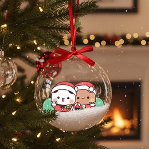 May include: A clear glass Christmas ornament with a red ribbon and bow. Inside, two cartoon figures in Santa hats stand on a bed of white snow. The ornament hangs on a decorated Christmas tree with lights.