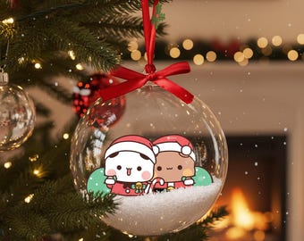 May include: A clear glass Christmas ornament with a red ribbon and bow. Inside, two cartoon figures in Santa hats stand on a bed of white snow. The ornament hangs on a decorated Christmas tree with lights.