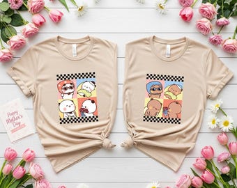 May include: Two beige t-shirts with cartoon animal graphics. Each shirt features a checkered border and a grid of colorful squares with different animal characters. The shirts are surrounded by pink flowers and greenery.
