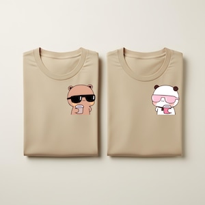 Può includere: Due magliette beige, ciascuna con una grafica a cartoni animati sul petto. Una presenta un orso marrone con occhiali da sole neri e una tazza di caffè. L'altra mostra un panda con occhiali da sole rosa e una tazza di caffè.