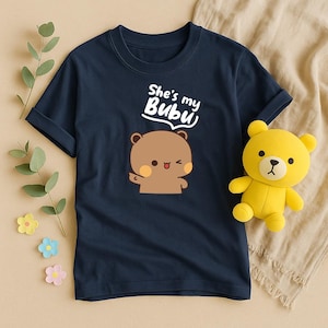 May include: Navy blue t-shirt featuring a cartoon bear with the text "She's my Bubu". A yellow teddy bear is next to the shirt. The shirt has a round neckline and short sleeves. The bear graphic is brown with a pink blush.