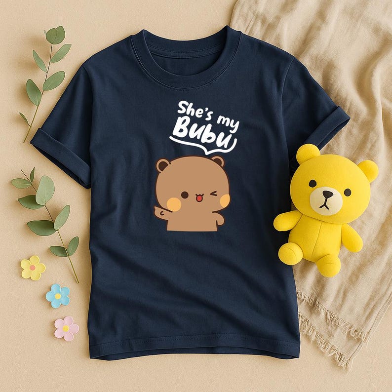 May include: Navy blue t-shirt featuring a cartoon bear with the text "She's my Bubu". A yellow teddy bear is next to the shirt. The shirt has a round neckline and short sleeves. The bear graphic is brown with a pink blush.