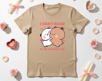 May include: A tan t-shirt featuring a cartoon illustration of two hugging bears with the text "I can't bear to be without you" in pink. The shirt is surrounded by pink roses, heart-shaped dishes, and candles.