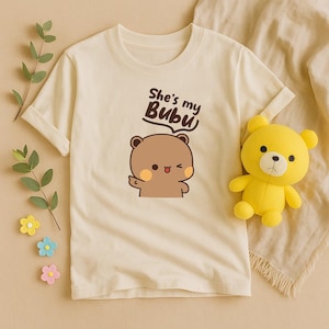 May include: Cream-colored t-shirt featuring a cartoon bear with the text "She's my Bubu". A yellow teddy bear is on the right side of the shirt. The shirt has rolled-up sleeves.