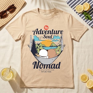 Peut inclure: T-shirt beige avec un motif de deux ours dans un hamac, avec le texte "The Adventure Soul Nomad". Le motif comprend un lac, des montagnes et un coucher de soleil. La phrase "The lake and mountain is calling nature made" est &eacute;galement imprim&eacute;e.