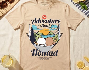 May include: Beige t-shirt with a graphic of two bears in a hammock, with the text "The Adventure Soul Nomad". The design includes a lake, mountains, and a sunset. The phrase "The lake and mountain is calling nature made" is also printed on the shirt.